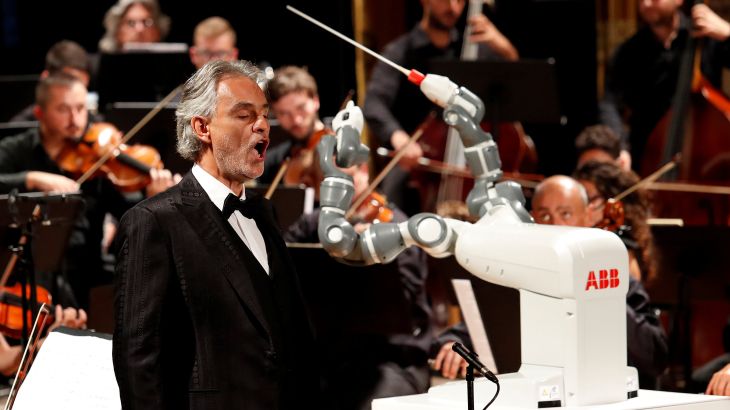 الروبوت (يومي) وهو يرفع عصاه ليقود أوركسترا لوكا فيلهارمونيك خلف التينور الإيطالي الشهير أندريا بوتشيلي